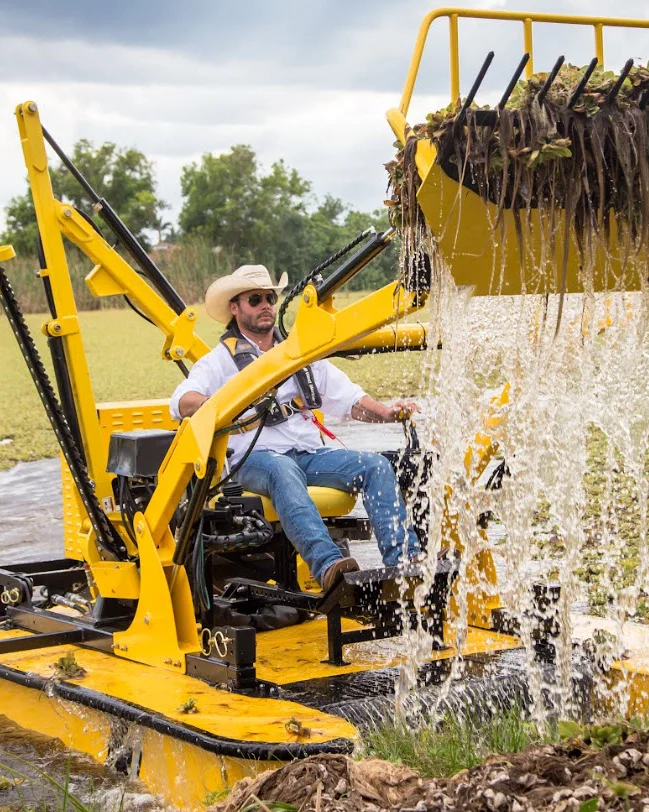 An operator lifting a full rake of vegetation and water out of a pond with a Weedoo workboat