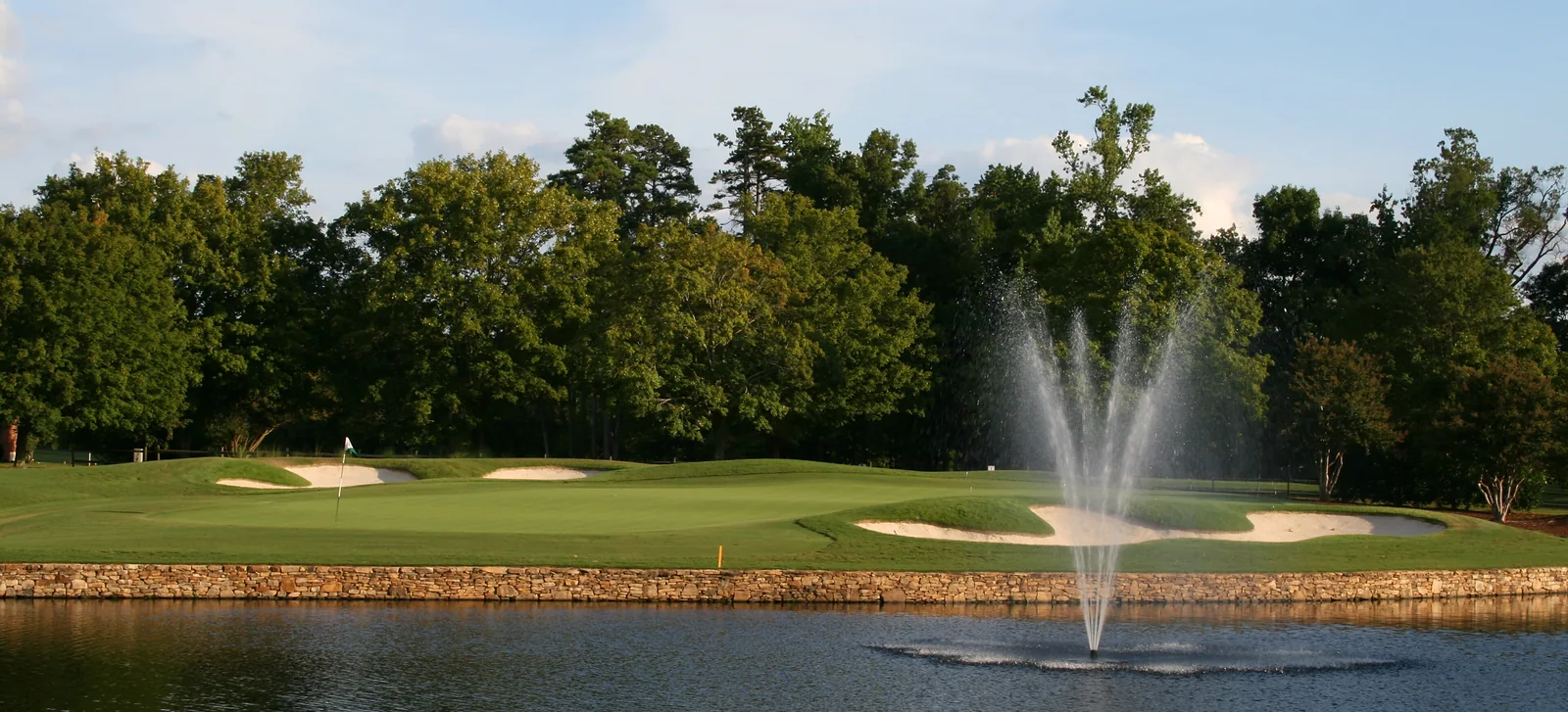 A fountain playing in a golf course pond with fairways and a clubhouse beyond