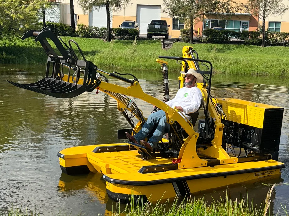 A Weedoo workboat clearing vegetation from a pond, its rake arm extended over the water